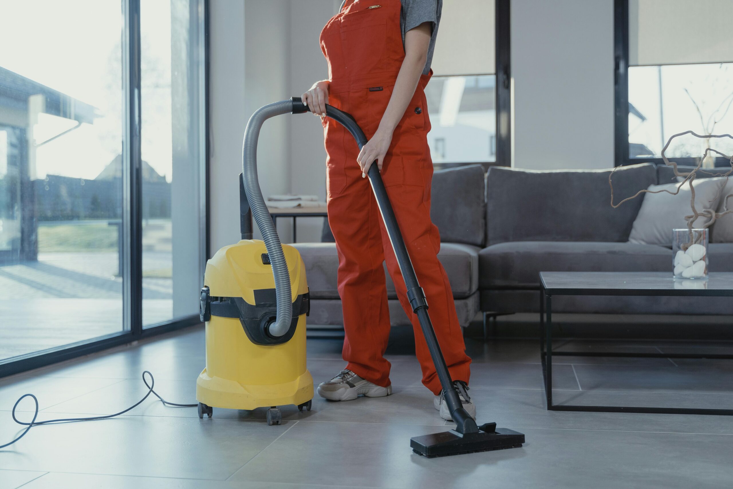 A professional cleaner in red coveralls vacuuming a modern living room with a yellow vacuum cleaner.