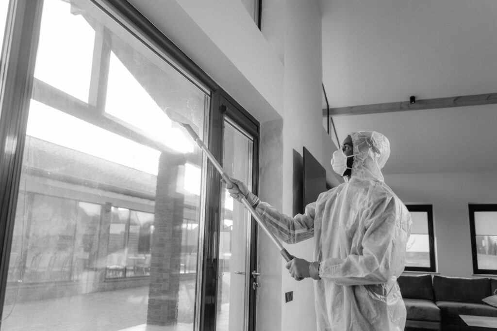 Person in protective coveralls sanitizing a window indoors with daylight.